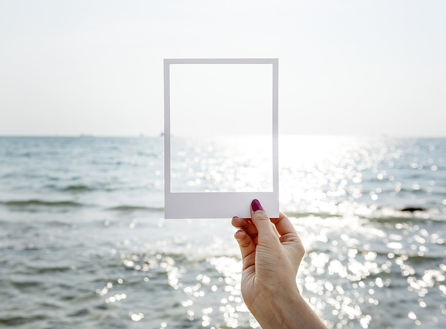 Hand holding blank polaroid frame against sparkling ocean background
