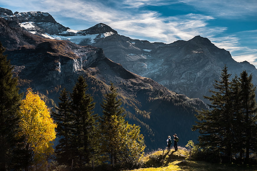 Hikers on Mountain Trail with Autumn Trees and Snowy Peaks
