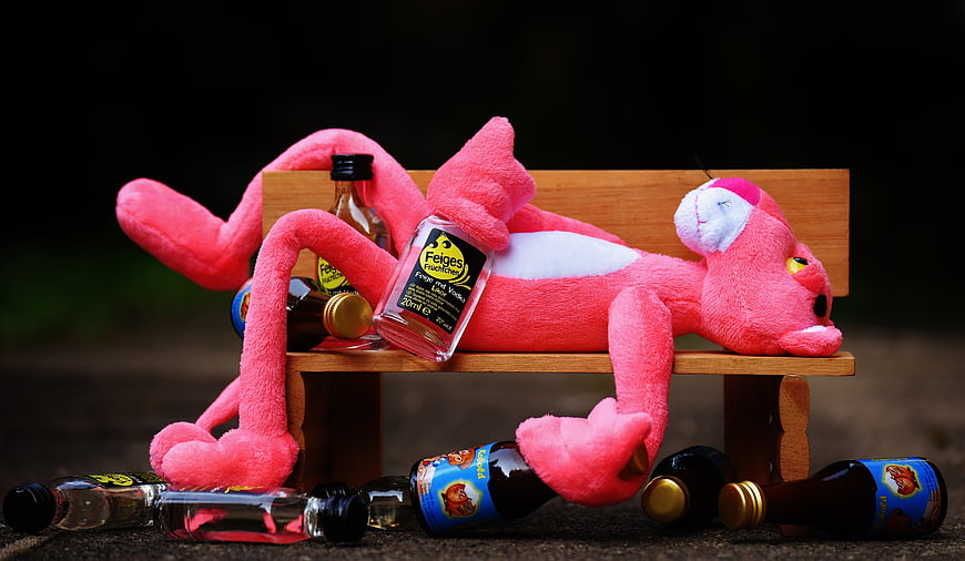 Pink Panther plush toy drunk on miniature bench with alcohol bottles
