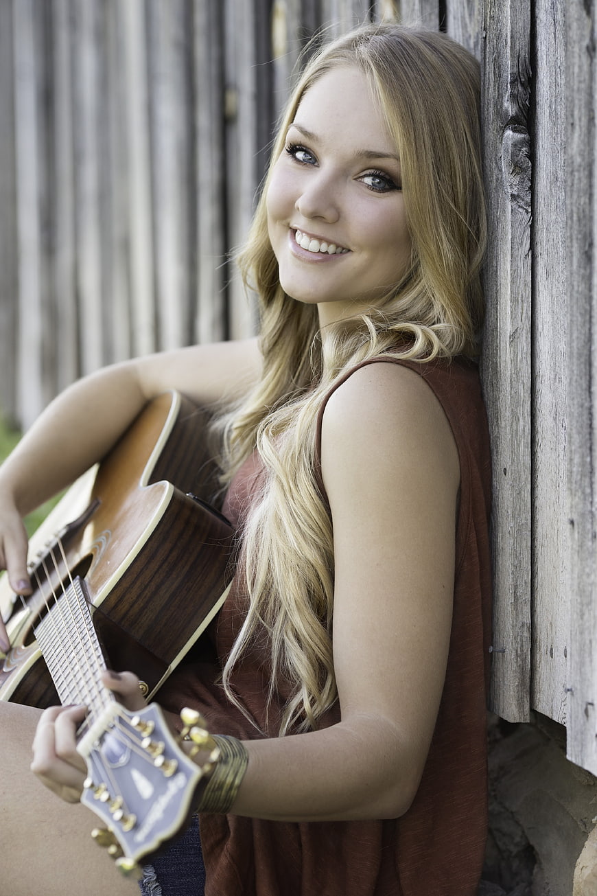 Smiling Blonde Woman Playing Acoustic Guitar by Wooden Fence
