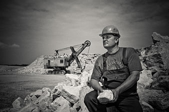 Miner in Hard Hat Sitting on Rocks with Excavator