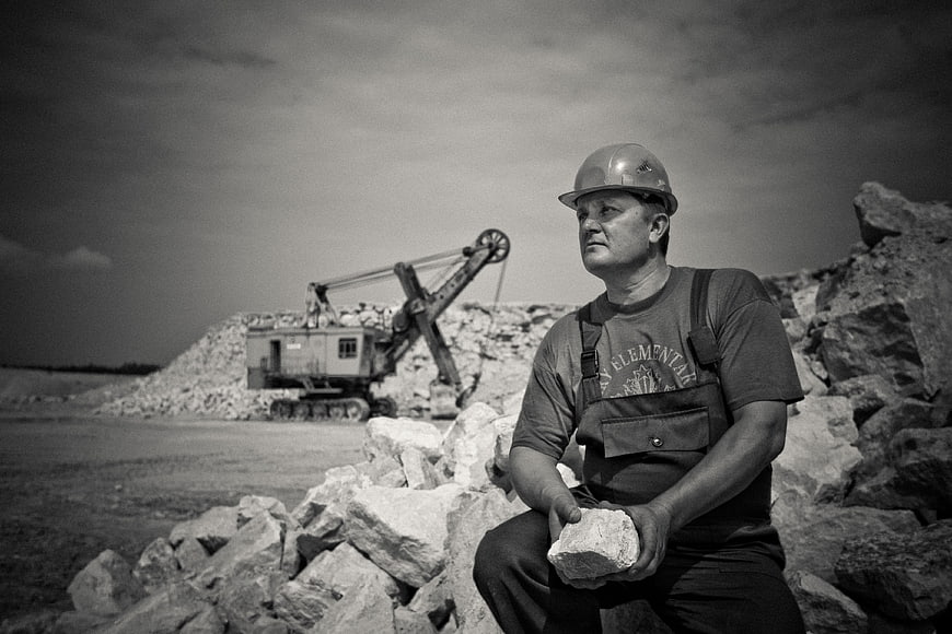 Miner in Hard Hat Sitting on Rocks with Excavator
