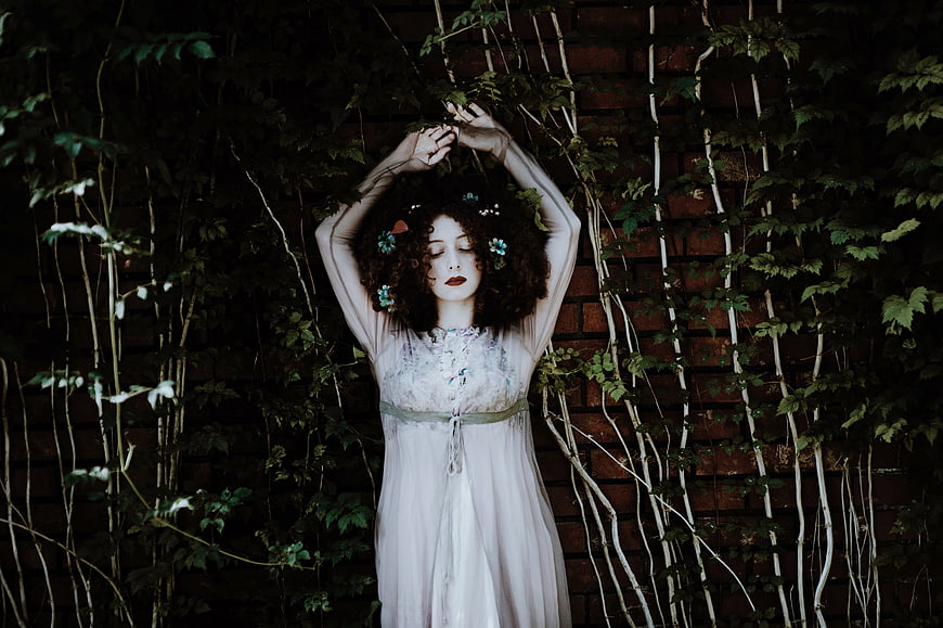 Woman in white dress with flower crown against ivy wall