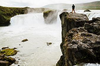 Person standing on cliff edge overlooking powerful waterfall