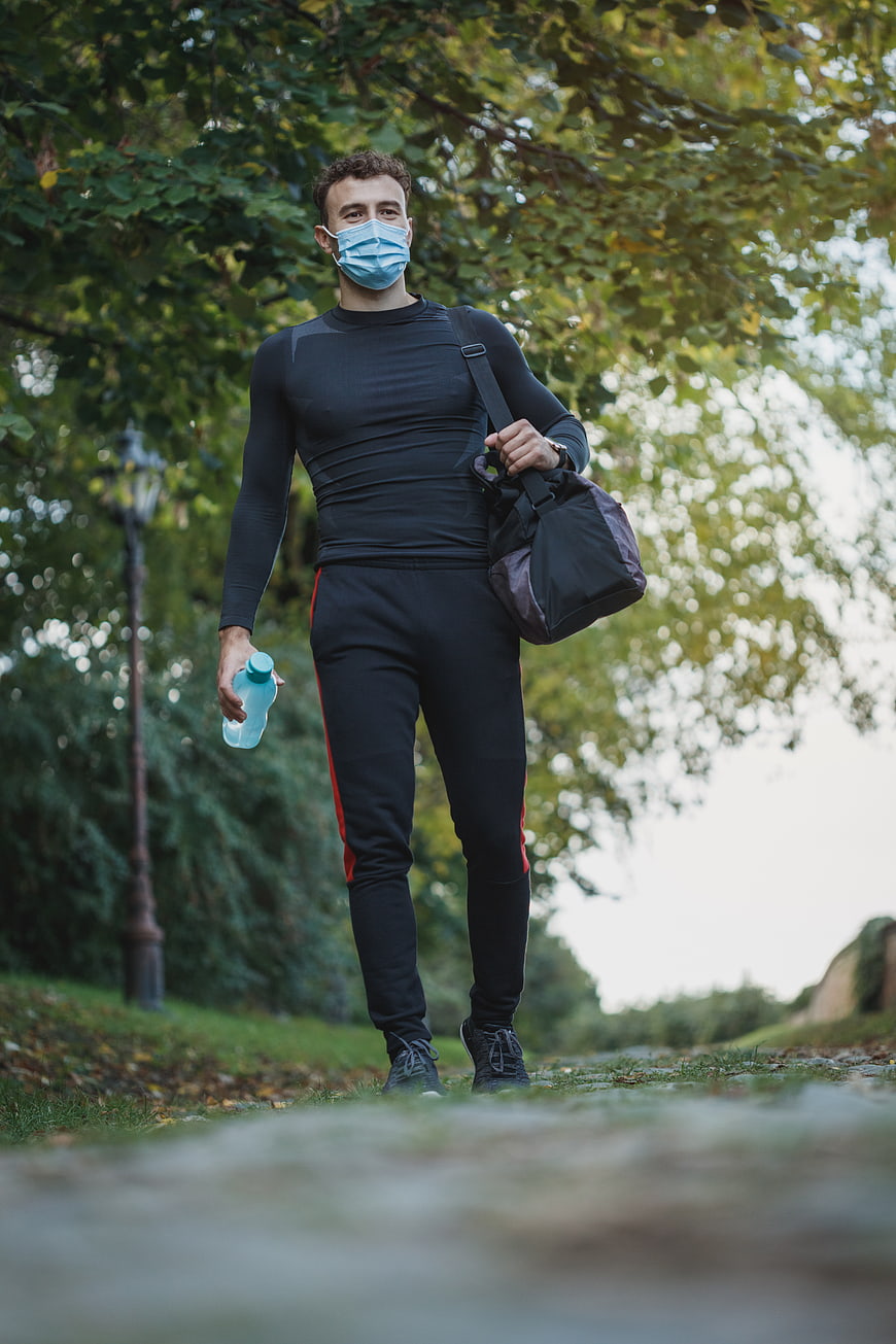 Athletic man with gym bag and mask walking in park