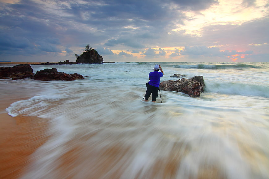 Photographer capturing sunset on rocky beach with long exposure waves