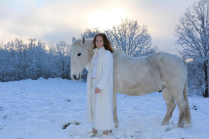 Woman in White Coat Standing with White Horse in Snow