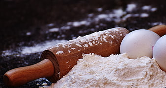 Wooden rolling pin, flour, and eggs on dark surface