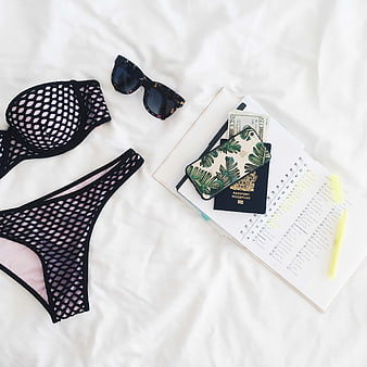 Flat lay of black mesh bikini, sunglasses, passport, and travel planner on white bed
