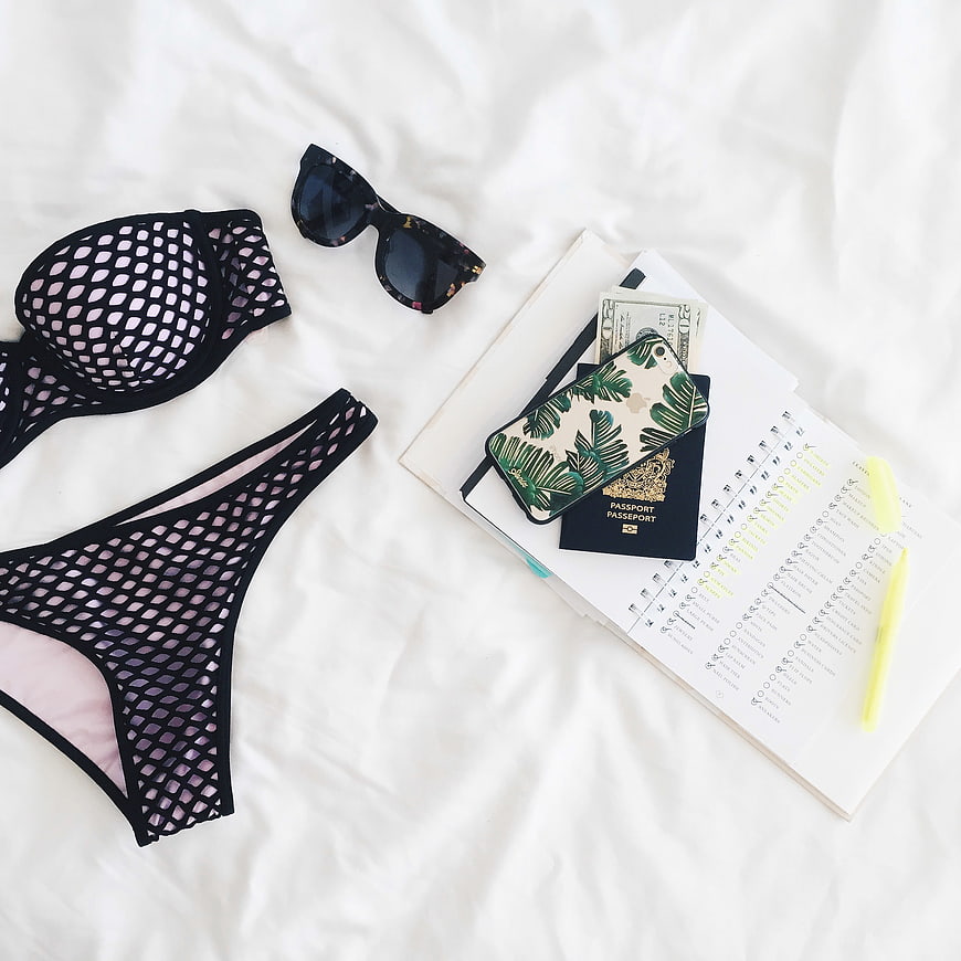 Flat lay of black mesh bikini, sunglasses, passport, and travel planner on white bed