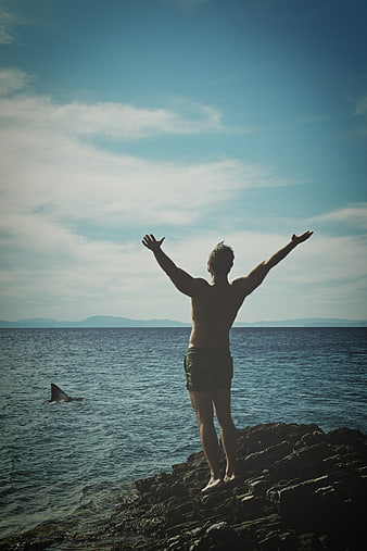 Man standing on rocks with arms raised near shark fin in ocean