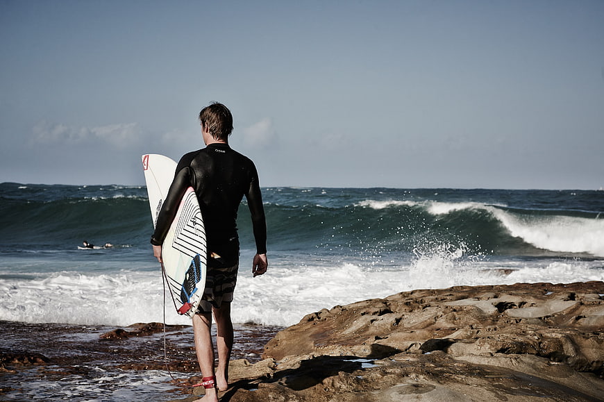 Surfer with board walking on rocky beach toward ocean waves