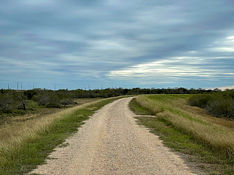 Winding gravel road through rural grassy landscape under cloudy sky
