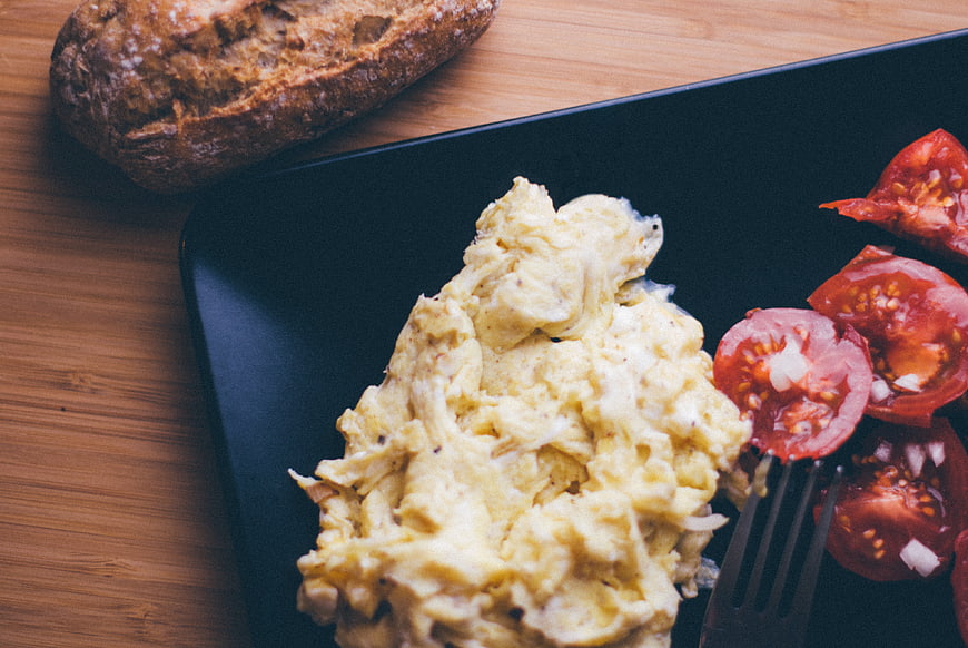 Overhead shot of scrambled eggs, tomatoes, and bread on a wooden table