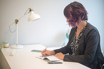 Woman with purple hair writing in notebook at white desk