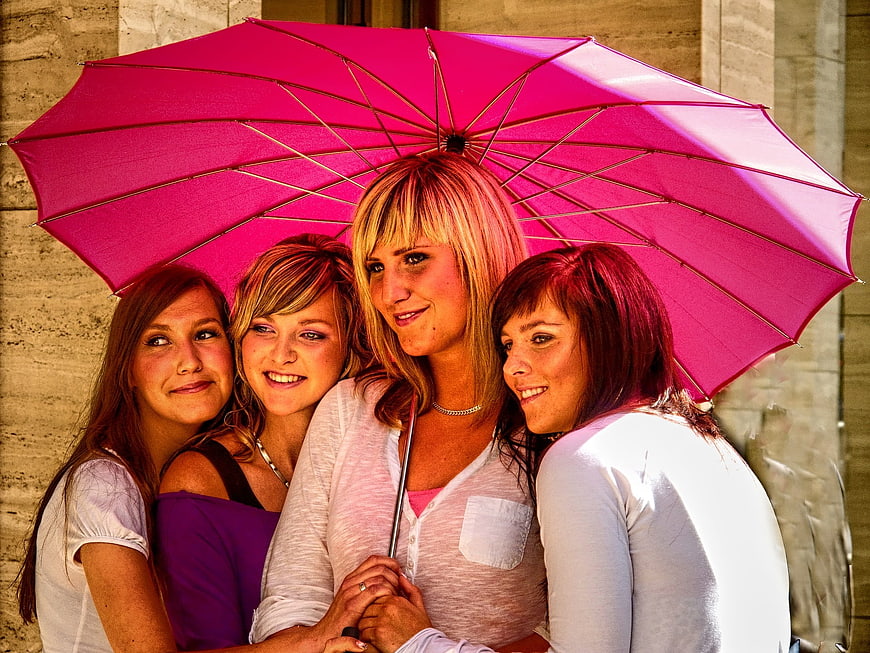 Four women smiling under a large pink umbrella