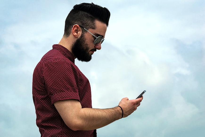 Bearded man in sunglasses using smartphone against cloudy sky