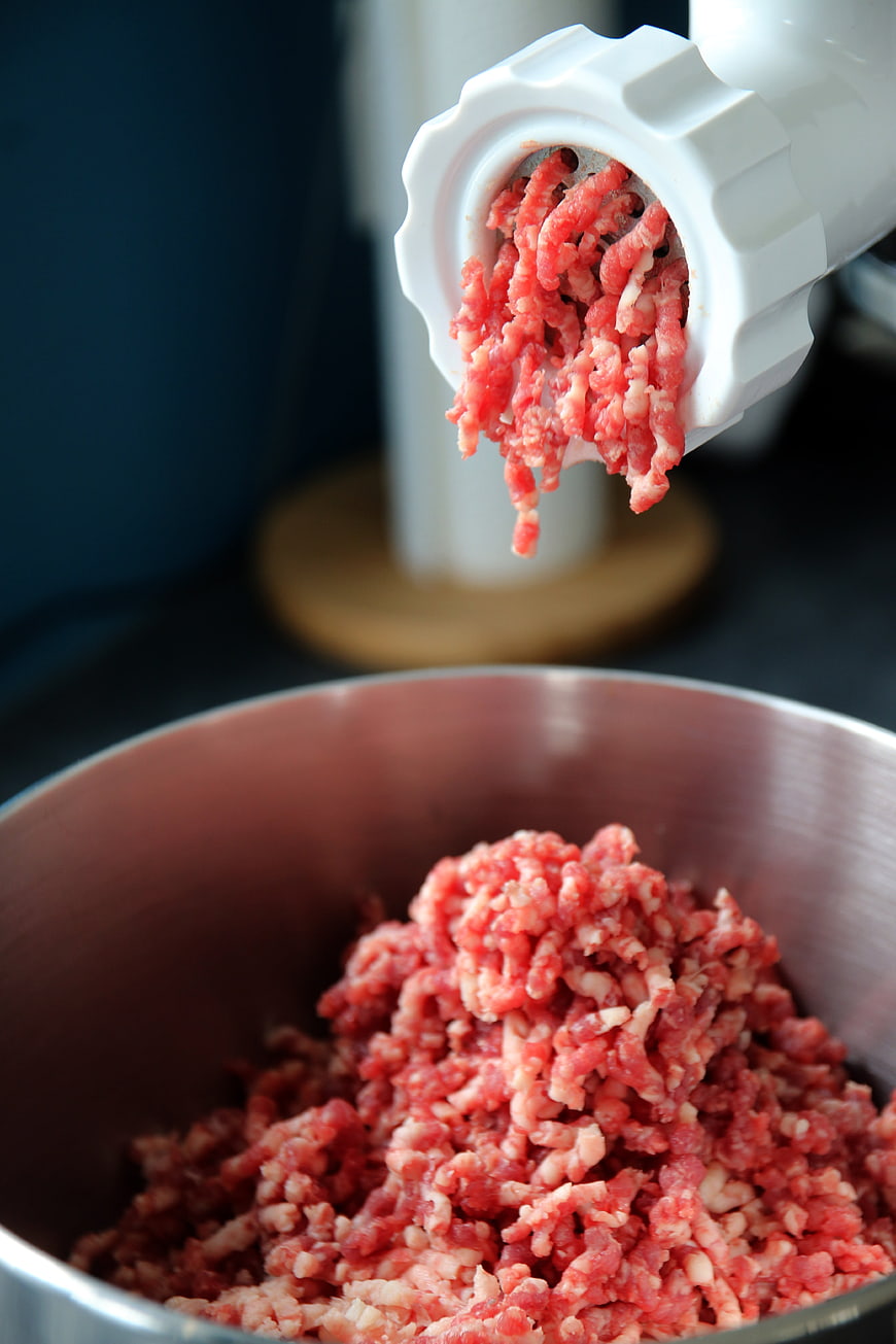 Fresh ground beef extruding from a white meat grinder into a metal bowl