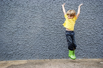 Happy boy jumping in green rain boots against gray wall