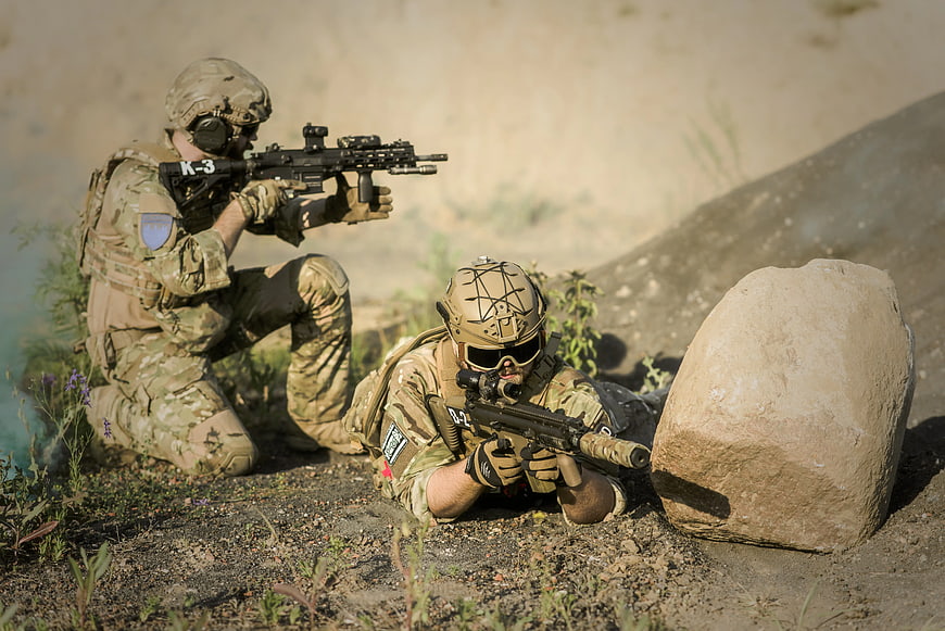 Two soldiers in tactical gear aiming rifles near a rock