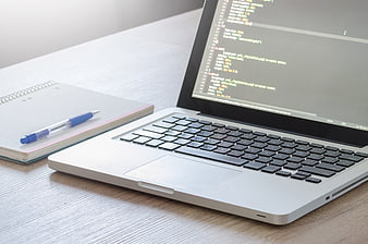 Laptop with code on screen and notebook on wooden desk