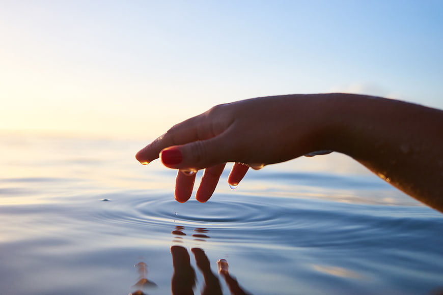 Woman's hand touching calm ocean water at sunset