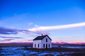Abandoned white house in snowy field with mountains at dusk