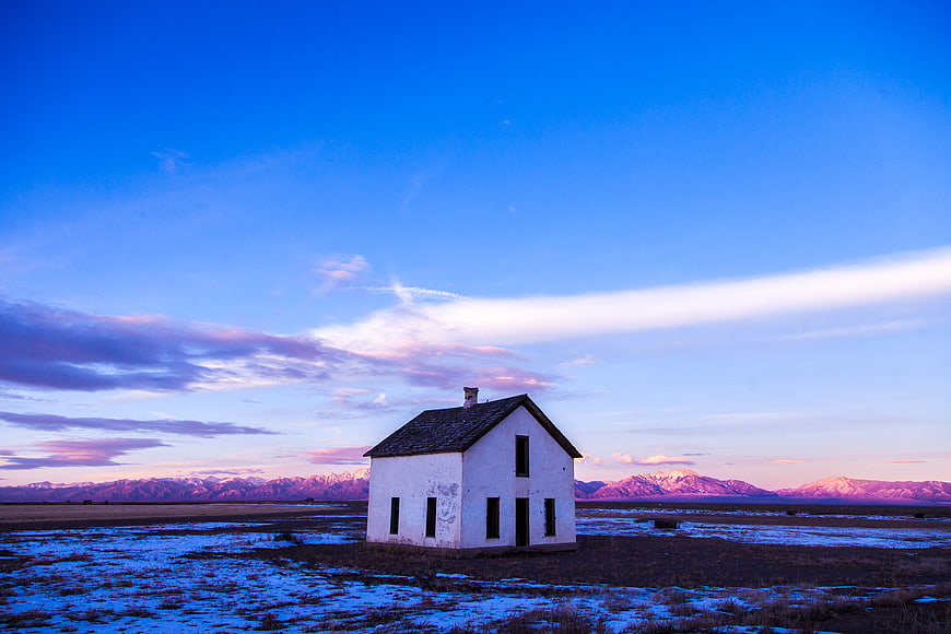 Abandoned white house in snowy field with mountains at dusk