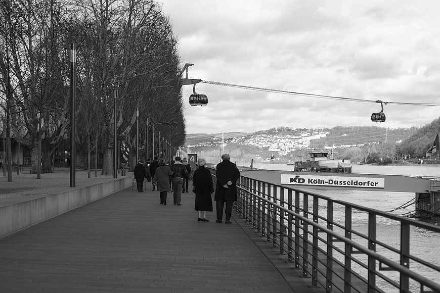 Black and white photo of people walking on a riverside promenade with cable cars