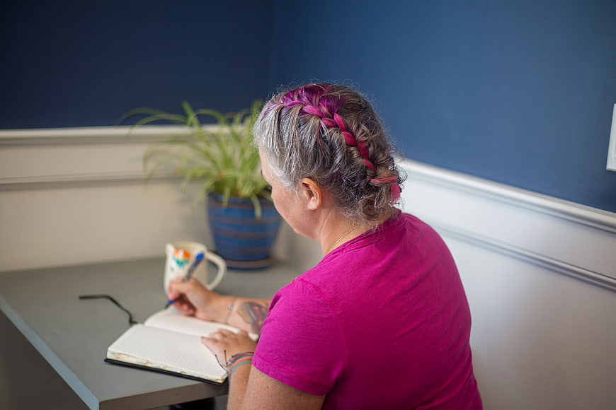 Woman with pink braided hair writing in a notebook at a desk