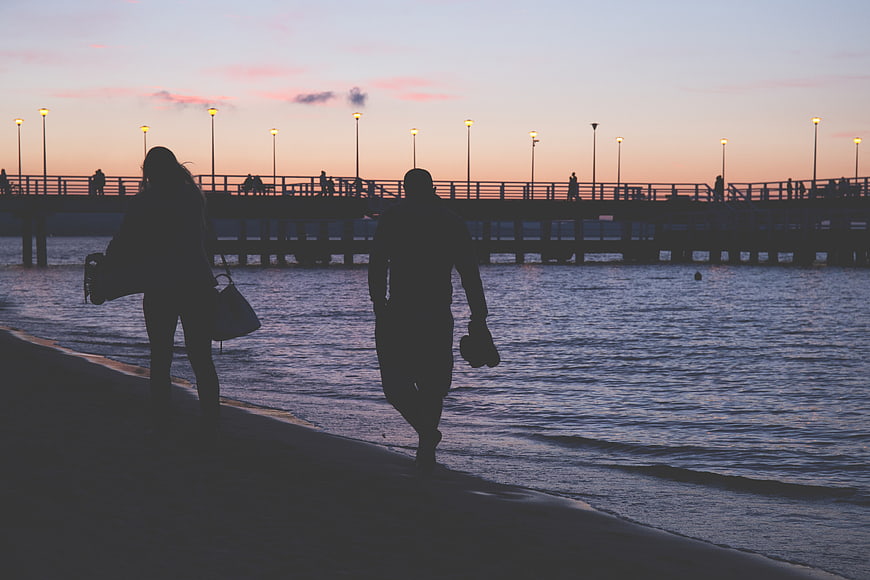 Silhouette of couple walking on beach at dusk with pier