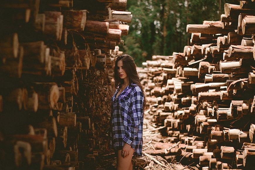 Woman in Plaid Shirt Standing Between Stacks of Logs