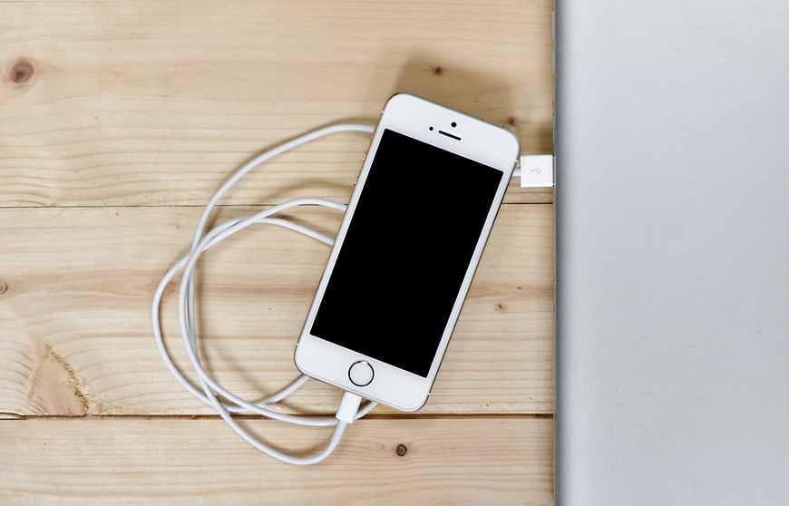 White smartphone charging via USB cable on wooden desk