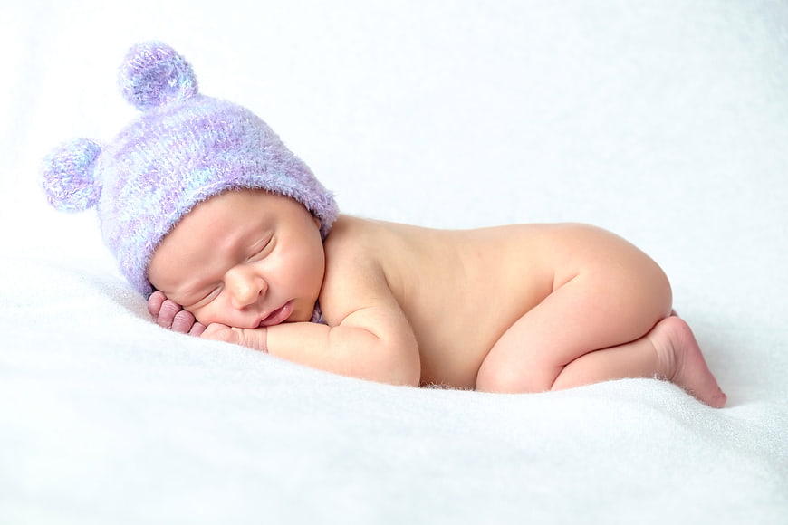 Sleeping newborn baby wearing purple knit hat on white blanket