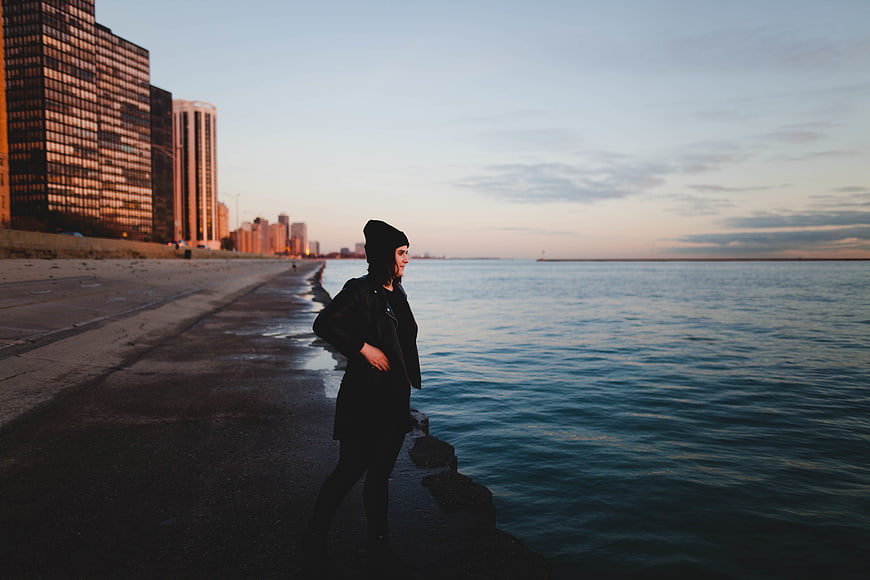 Woman in black coat standing by city waterfront at sunset