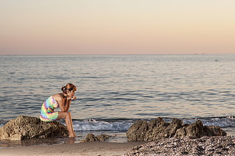 Contemplative Woman in Colorful Dress Sitting on Beach Rocks at Sunset