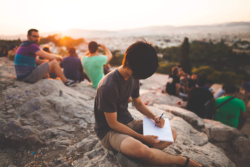 Young man sketching on rocks overlooking city at sunset