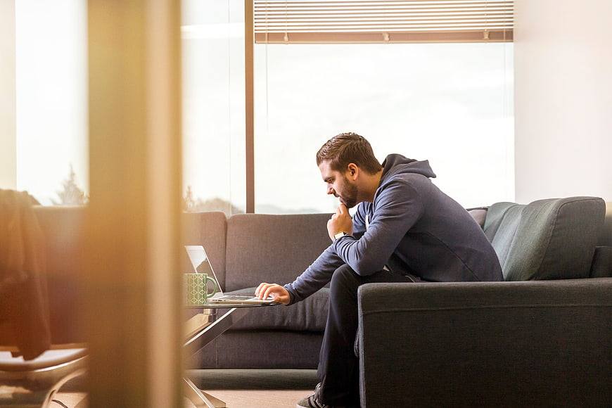 Thoughtful man working on laptop on sofa in bright living room