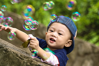 Toddler in denim hat reaching for colorful soap bubbles in a park