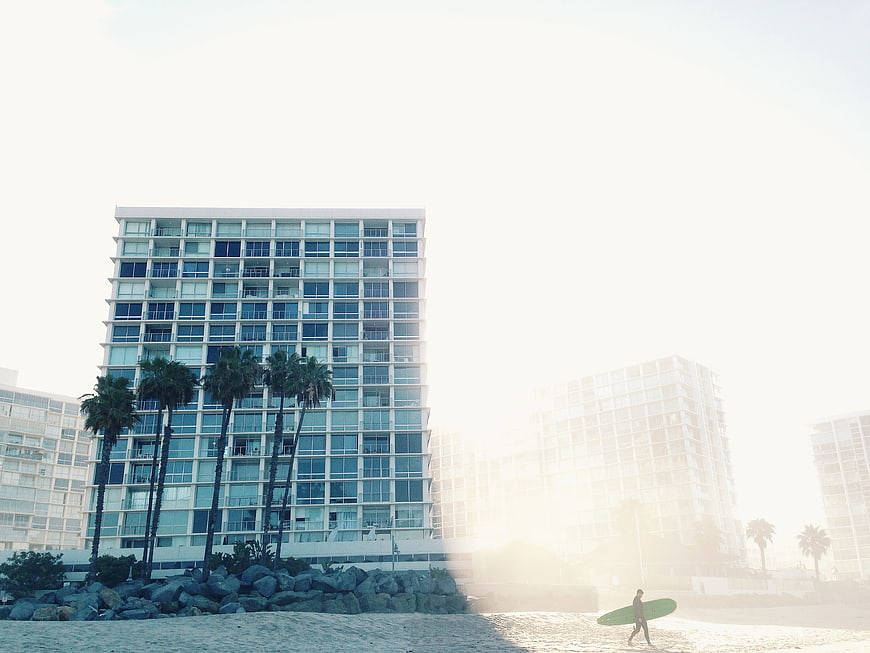 Surfer walking on beach with modern high-rise buildings and palm trees