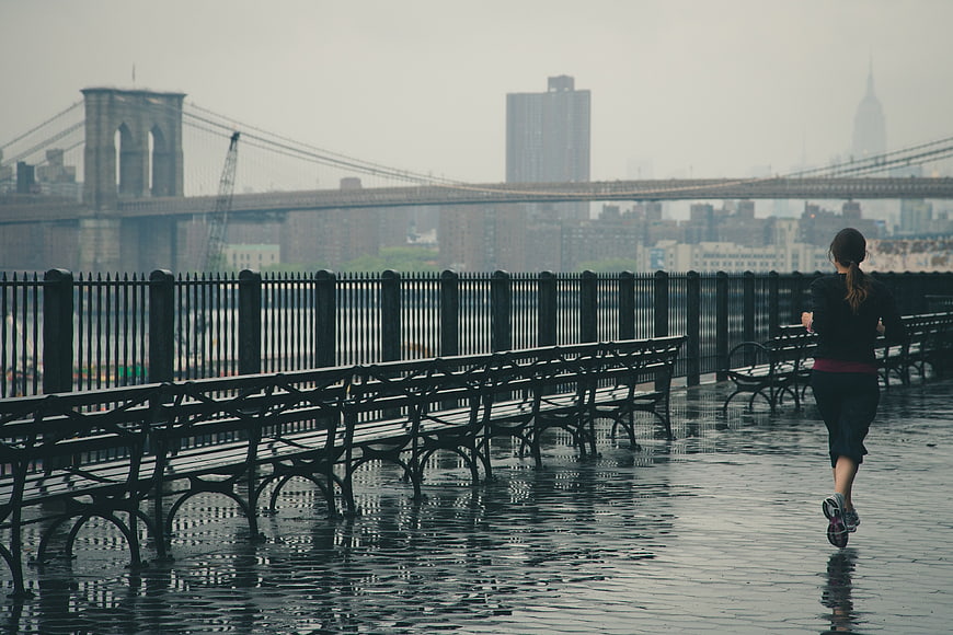 Woman jogging on wet promenade with Brooklyn Bridge background