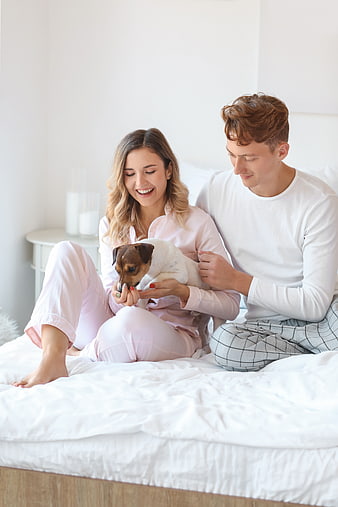 Happy couple in pajamas playing with small dog on bed