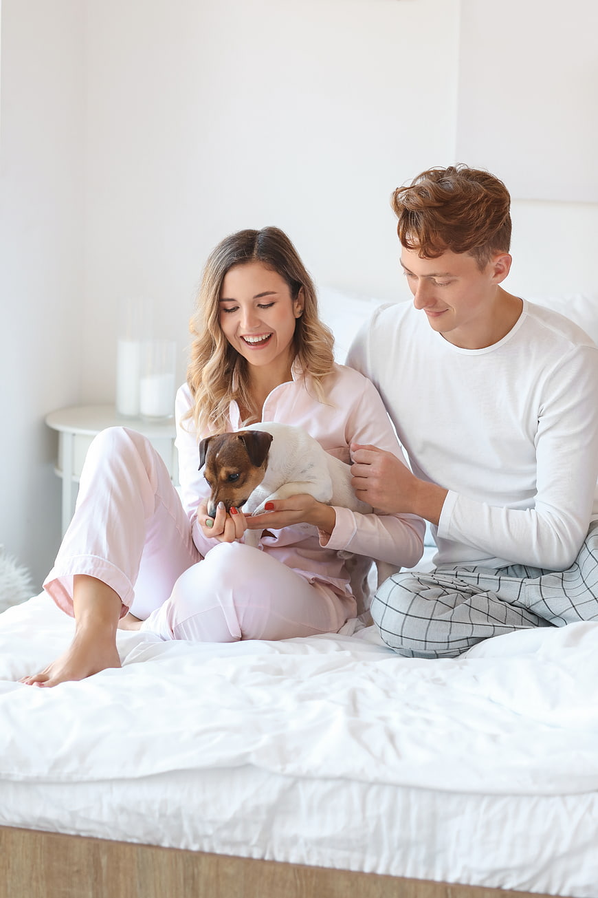 Happy couple in pajamas playing with small dog on bed