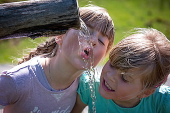 Two young girls drinking water from a wooden spout outdoors