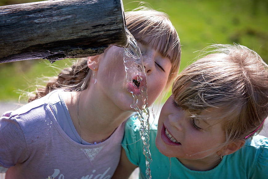 Two young girls drinking water from a wooden spout outdoors