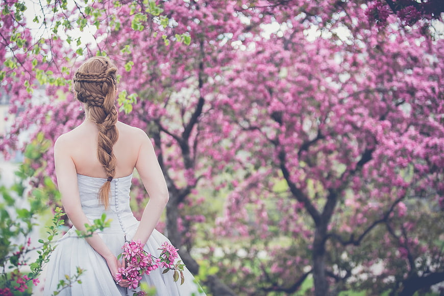 Bride with Braided Hair Holding Pink Flowers Under Blossoming Tree