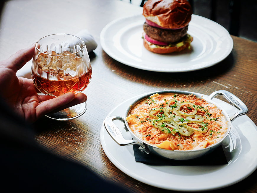 Hand holding whiskey glass with burger and mac and cheese on wooden table