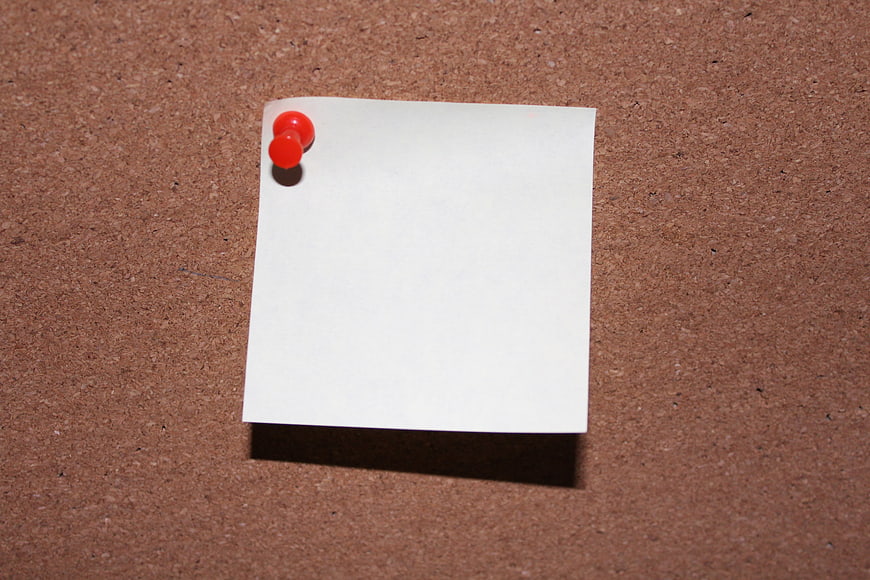 Blank white sticky note pinned to corkboard with red pushpin