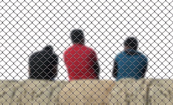 Three blurred men behind a chain-link fence in a camp