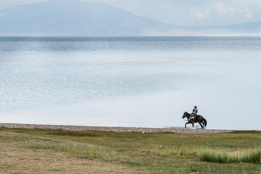 Man Riding Horse Along Lake Shore with Mountains
